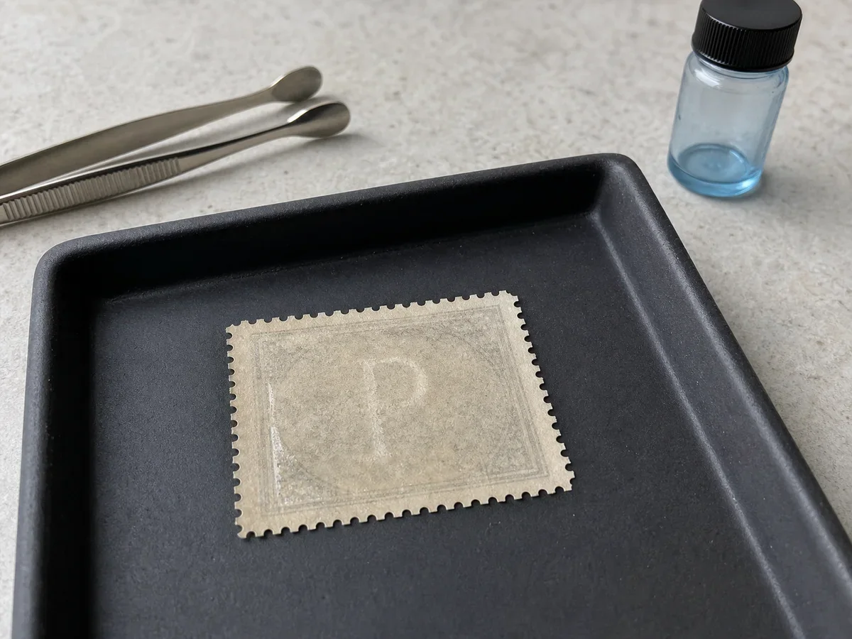Fictional stamp lying face down in a matte black tray with tongs and a capped unbranded watermark-fluid bottle nearby